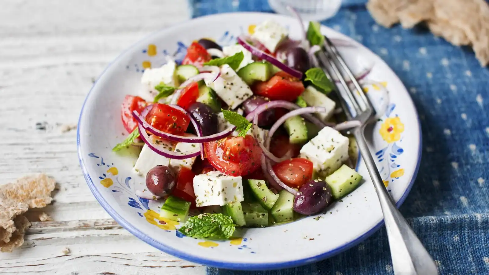 A plate of Greek salad with tomatoes, cucumbers, olives, red onions, feta cheese, and fresh herbs, served on a floral plate with a fork.