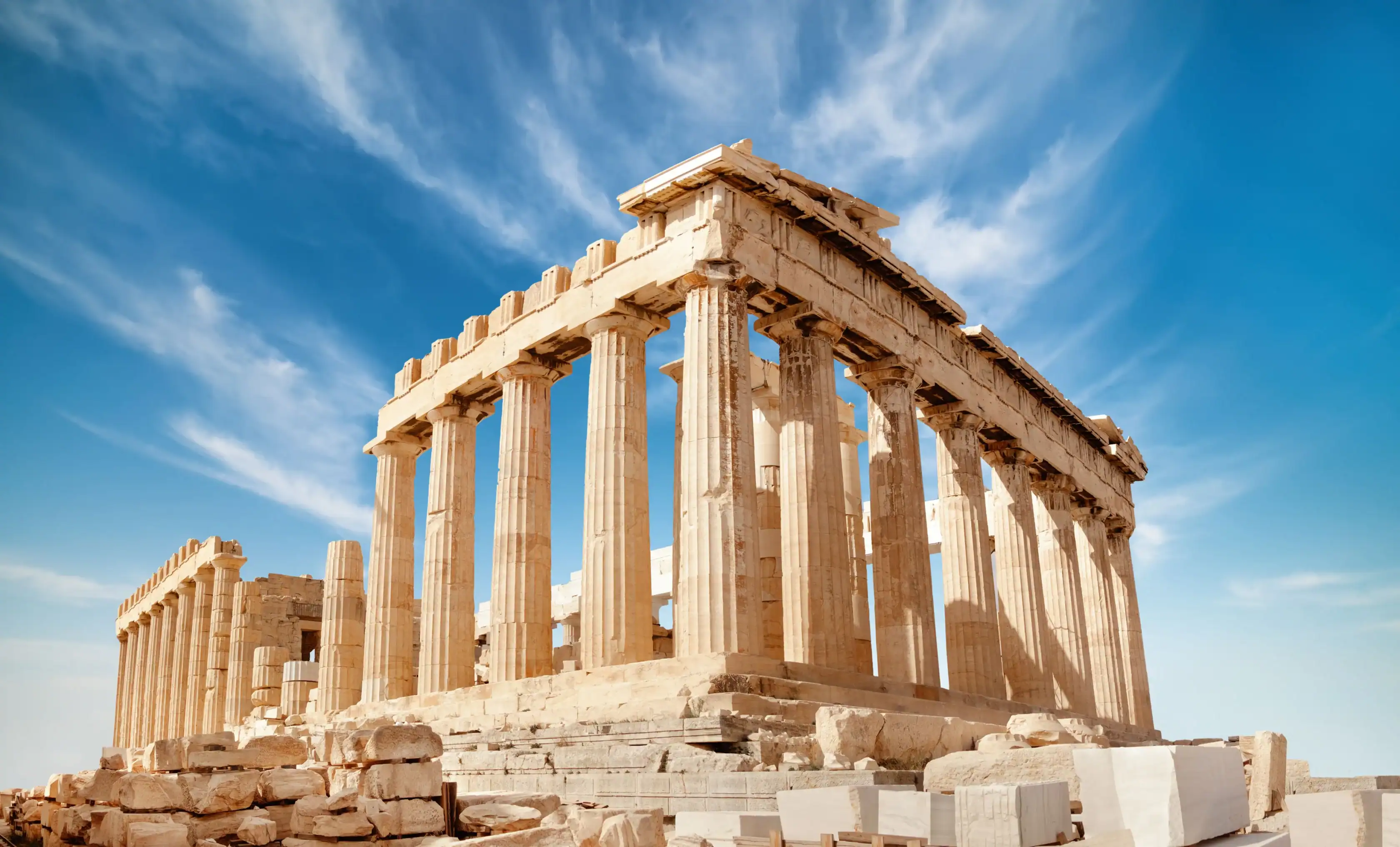 The Parthenon, an ancient Greek temple with columns and partial ruins, stands under a blue sky with wispy clouds.