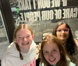 Three girls smile and pose indoors near a window with bold white text printed on the glass, while a street scene is visible outside at night.