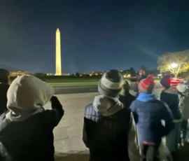 A group of people, some in winter clothing, stand at night facing the illuminated Washington Monument in the distance.