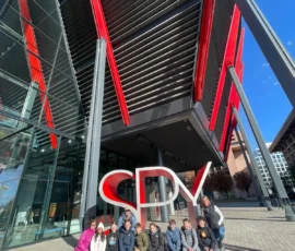 A group of people sit in front of a large “SPY” sign outside a modern building with red and gray architectural features.