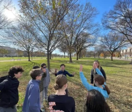 A group of eight people stand in a circle on a grassy field with leafless trees, engaging in conversation under a clear blue sky.