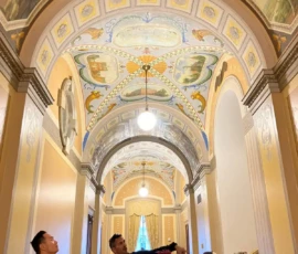 A group of people stand in a hallway with ornate, colorful murals and decorative arches on the ceiling, admiring the artwork above.