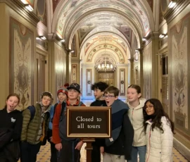 A group of young people stand in front of a "Closed to all tours" sign inside an ornate, arched hallway with decorative ceilings and patterned floors.