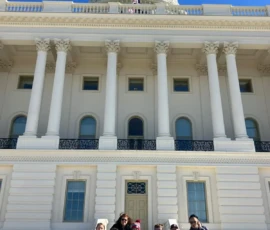 A group of people are sitting on the steps in front of the U.S. Capitol building under a clear blue sky.