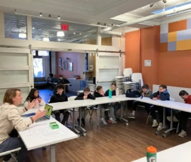 A teacher sits at a table presenting to a group of students, who are seated in a U-shaped arrangement in a classroom with orange walls and stackable chairs.