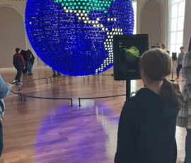 A girl stands in front of an interactive screen with a large digital globe display in a museum gallery; visitors are seen in the background.