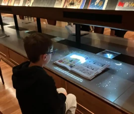 A boy sits at a library table looking at an open interactive book, which is illuminated with projected lights and information. Books are displayed on shelves in the background.