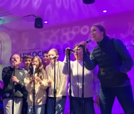 A group of six people stand together singing into microphones on a purple-lit stage, while another person sits in the background watching.