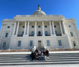 A group of people sits on the front steps of a large white neoclassical building with columns and a dome under a clear blue sky.