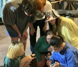 Six people, mostly in casual clothes and sandals, gather closely on a wooden deck at night, looking at a smartphone held by one person sitting on the ground.