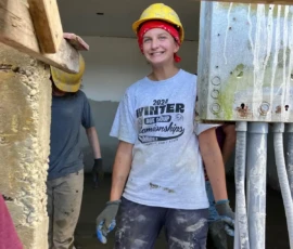A person wearing a yellow hard hat, red bandana, and work gloves stands in a partially constructed building with others, smiling and covered in dust and paint.