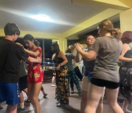 A group of young people dance together indoors under fluorescent lighting, with pairs holding hands and others moving in the background.