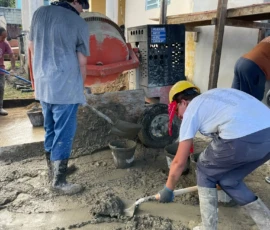 Two people wearing helmets and boots spread wet cement on the ground at a construction site, with a cement mixer and various tools visible nearby.