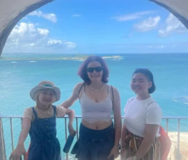 Three people stand smiling in front of a stone archway with a view of the blue ocean and cloudy sky in the background.