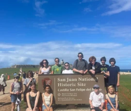 A group of people pose in front of the San Juan National Historic Site sign at Castillo San Felipe del Morro on a sunny day.