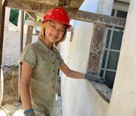 A person wearing a red hard hat, gloves, and a light-colored jumpsuit is smiling and working at a construction site next to a building.