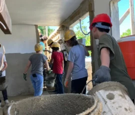 A group of people wearing hard hats and gloves carry buckets of cement inside a partially constructed building.