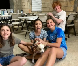 Four teenagers sit on the floor indoors, smiling and petting a small dog, with folding tables and chairs in the background.