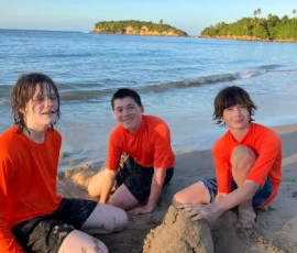 Three boys in orange shirts sit on a sandy beach building a sand structure, with the ocean and a green island visible in the background.