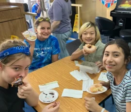 Four kids sit around a table in a casual restaurant, eating donuts and smiling at the camera. A woman walks in the background near a restroom sign and wall posters.