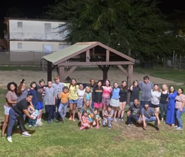 A large group of people, including children and adults, pose together at night under a wooden shelter on a grassy area.