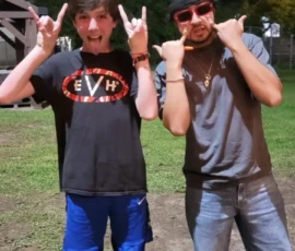 Two people stand outside at night on grass, both making hand gestures and posing playfully for the camera. One wears a black T-shirt and shorts, the other sunglasses, a hat, and jeans.