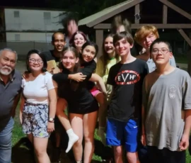 A group of thirteen people, both adults and teens, pose together outside at night in front of a wooden structure, smiling at the camera.
