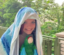 A smiling person stands outside with a towel draped over their head, holding a green parrot plush toy; lush greenery is visible in the background.