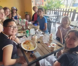 A group of nine young people sit around a table at an outdoor restaurant, eating and smiling at the camera. Plates of food and drinks are on the table.