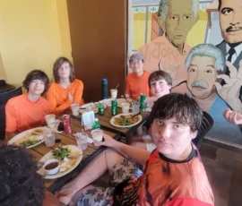 A group of young people sit around a table with food and drinks, next to a mural featuring two men and the words "TOMA Y DAMA" on the wall behind them.