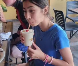 A girl in a blue shirt and denim shorts sits at an outdoor table, sipping a drink with a red straw. Other people are seated nearby.