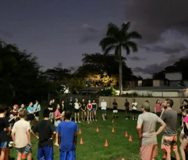 A group of people stand in a circle around orange cones on a grassy lawn at dusk, with palm trees and buildings in the background.
