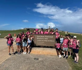 A group of people in pink shirts pose in front of the San Juan National Historic Site sign at Castillo San Felipe del Morro on a sunny day.
