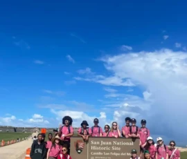 A group of people wearing pink shirts pose in front of a sign for San Juan National Historic Site, Castillo San Felipe del Morro, under a bright blue sky.