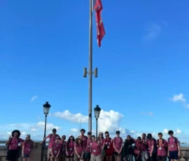 A group of people wearing matching pink shirts stands in front of a large flagpole with a flag, under a blue sky with clouds.