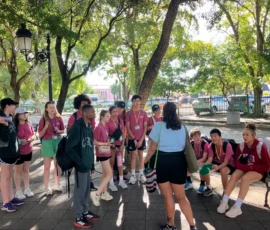 A group of students in matching maroon shirts listens to an adult leader outdoors under trees near a metal fence and urban buildings.