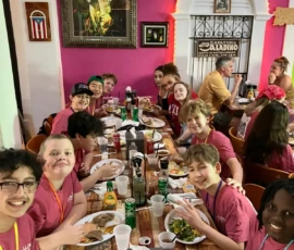 A group of kids and adults sit at a long table in a restaurant, eating a meal together. Most are wearing matching pink t-shirts, and the table is filled with food and drinks.