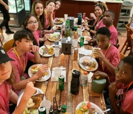 A group of young people in matching maroon shirts sit around a table eating a meal together in a restaurant, with a server visible behind the counter.