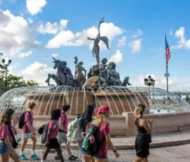 A group of people walk past the Raíces Fountain, featuring bronze statues and water jets, with a U.S. flag and blue sky in the background.