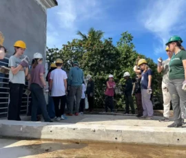 A group of people wearing hard hats and gloves stand on a construction site outdoors, some holding water bottles, with trees and blue sky in the background.