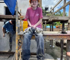A young person in a yellow hard hat, red shirt, and work gloves sits on scaffolding at a construction site, with building materials and blue sky in the background.