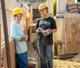 Two young people wearing hard hats and gloves stand at a construction site, with cement bags and buckets of rocks nearby.