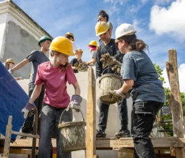 A group of people wearing hard hats and gloves work together on scaffolding, passing buckets of cement or mortar at a construction site under a partly cloudy sky.