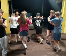 A group of teenagers practice partner dancing indoors at night, wearing casual clothes and forming pairs on a concrete floor.