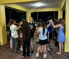 A group of teenagers stand in pairs under a yellow-roofed outdoor area at night, engaged in an activity that involves holding hands.