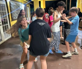 A group of teenagers are dancing and socializing in a brightly lit, covered outdoor area with yellow walls at night.