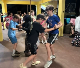 A group of kids dance in pairs in a covered outdoor area, with yellow pillars and casual clothing, during what appears to be a social event at night.