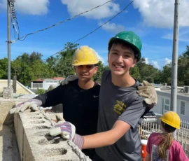 Two teens wearing construction helmets and gloves smile while building a concrete block wall outdoors; another person works in the background under a blue sky.