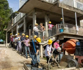 A group of people wearing yellow hard hats form a line to pass buckets of cement outside a concrete building under construction.